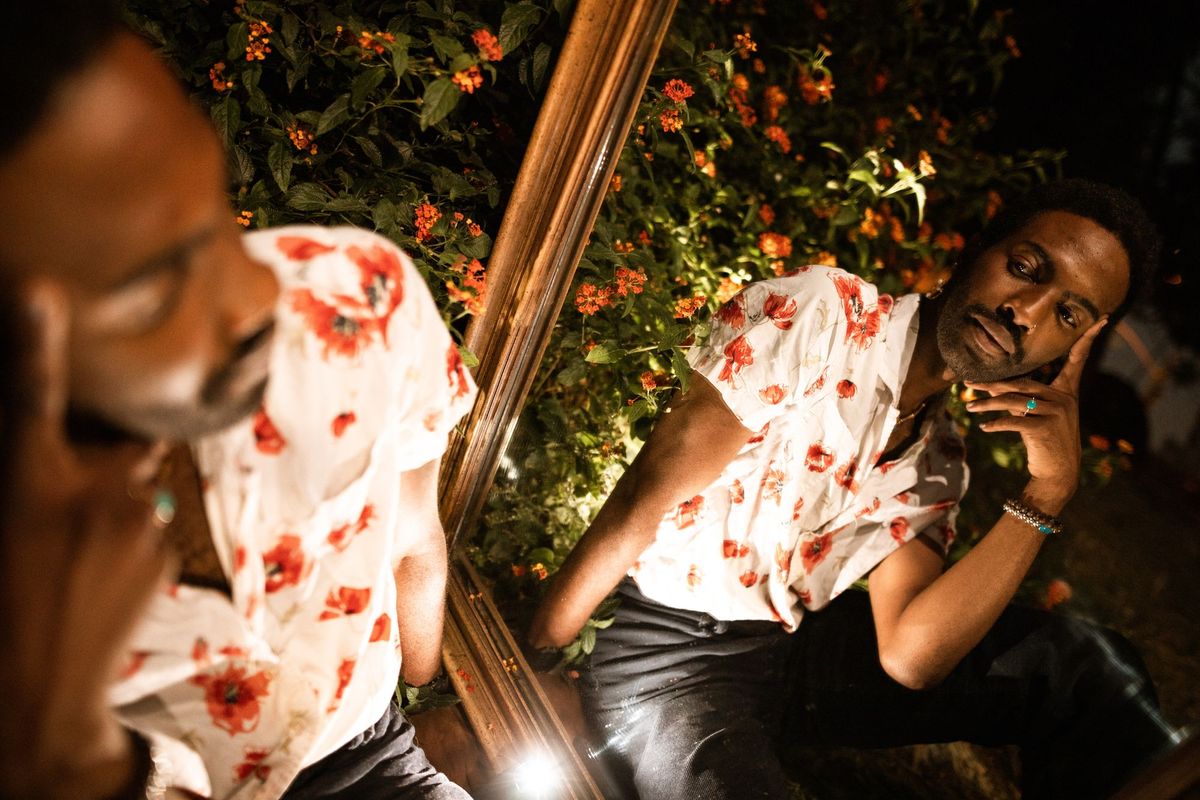 A Black man seated next to a mirror looks pensively at his reflection